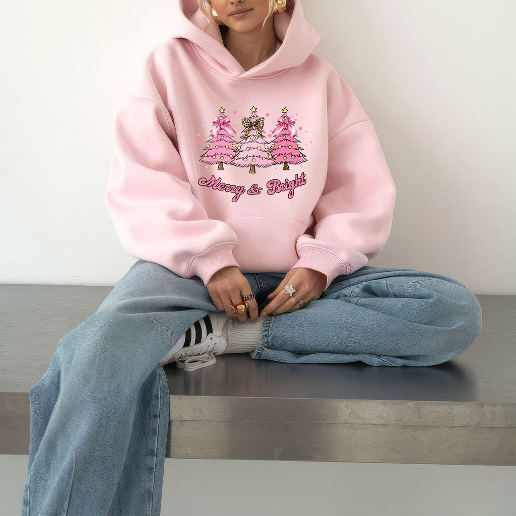 Woman wearing a pink hoodie with festive Christmas Trees and text, sitting on a bench.