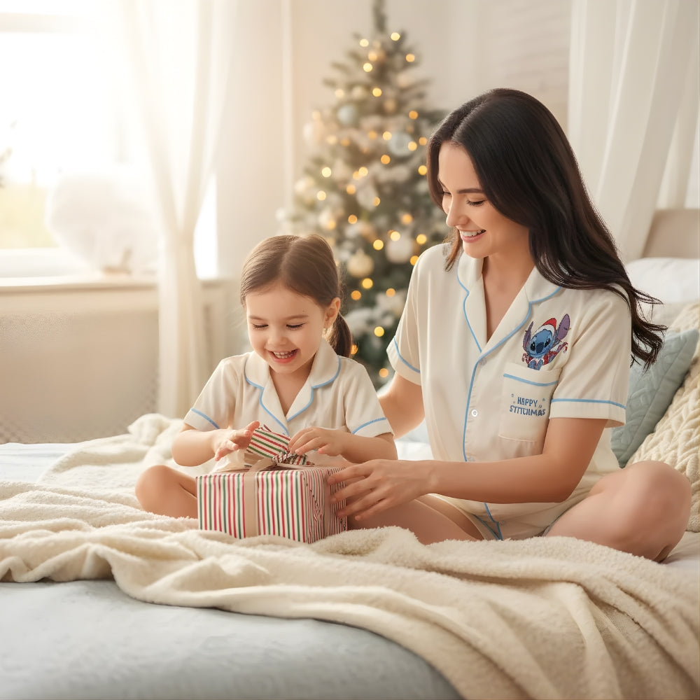 Woman and child in matching beige pajama set with Stitch in a Santa Claus outfit opening a gift in a cozy room with a Christmas tree.