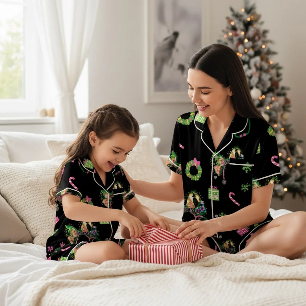 Woman and child in matching black Christmas-themed pajama set with Wicked designs opening a gift on a bed with a Christmas tree in the background
