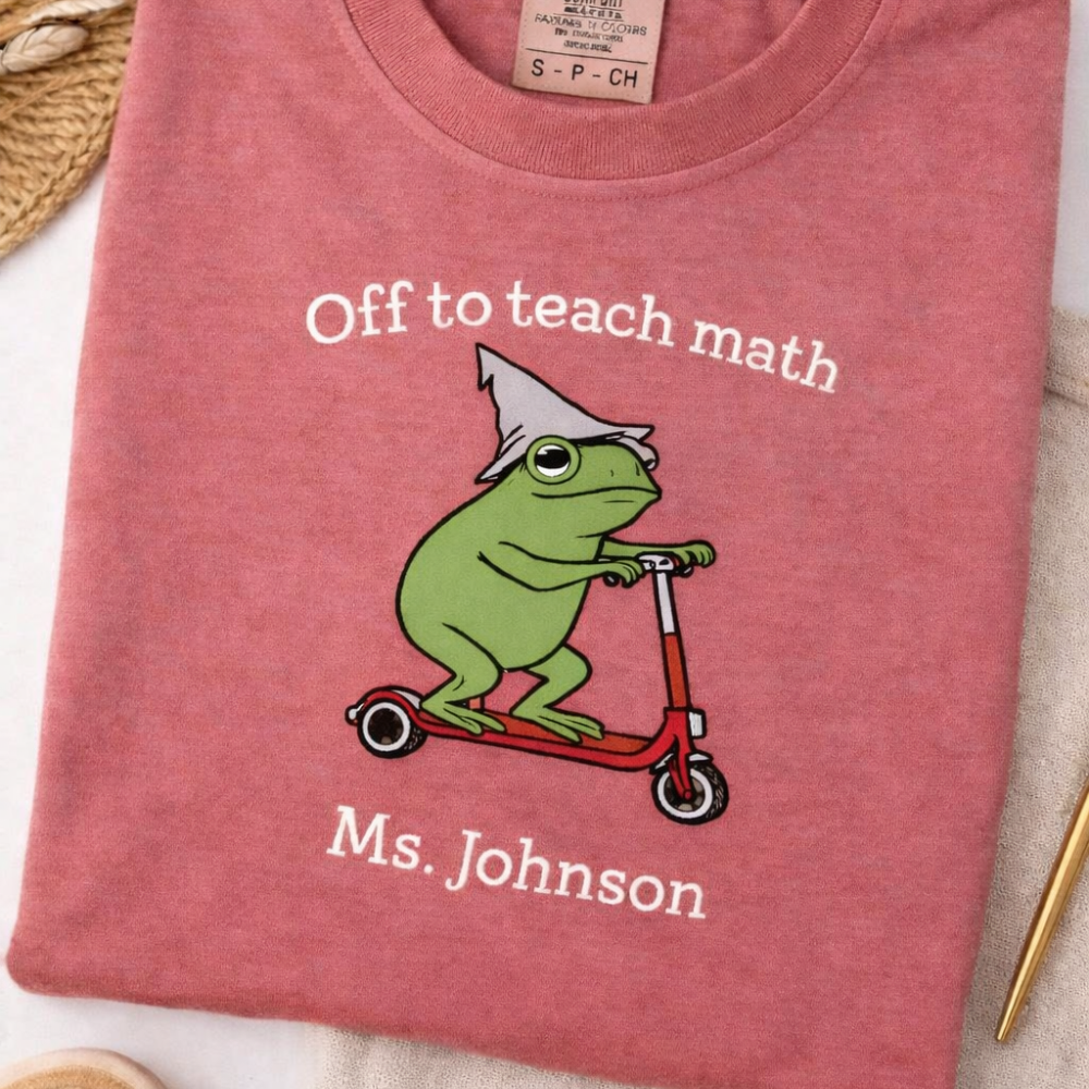 Pink t-shirt with a frog on a scooter graphic and text 'Off to teach math Ms. Johnson' on a light background.