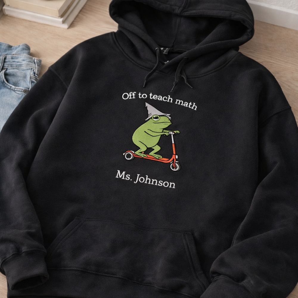 Black hoodie with a frog on a scooter design and text 'Off to teach math Ms. Johnson' on a wooden floor.